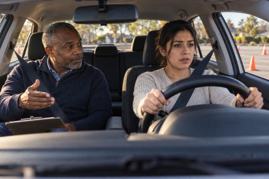 An adult driving instructor giving advice to a focused young female driver holding the steering wheel during a lesson