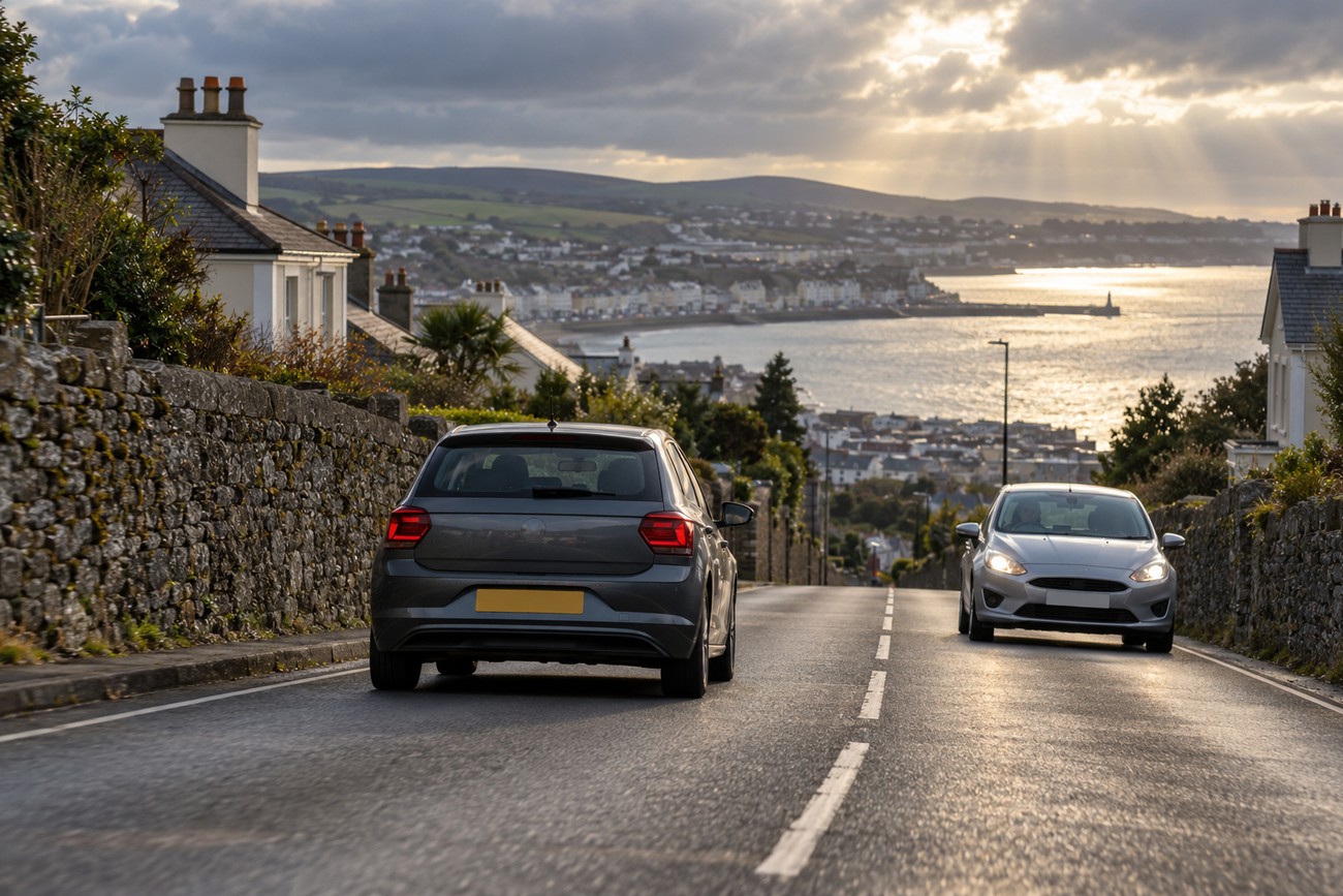 A grey VW Polo driving down a narrow coastal road flanked by stone walls with a town and sea view ahead