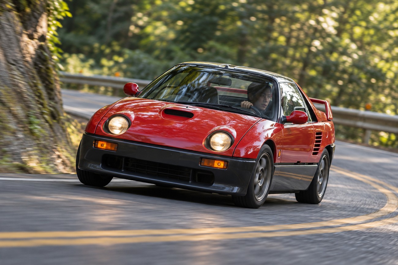 Red Autozam AZ-1 kei sports car navigating a sharp curve on a scenic mountain road with lush green trees