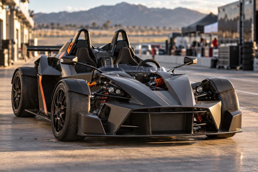Front three-quarter view of a dark carbon fiber KTM X-Bow sports car parked in a racing paddock during daytime
