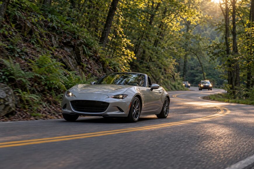 Silver Mazda Miata sports car driving on a winding mountain road surrounded by dense forest