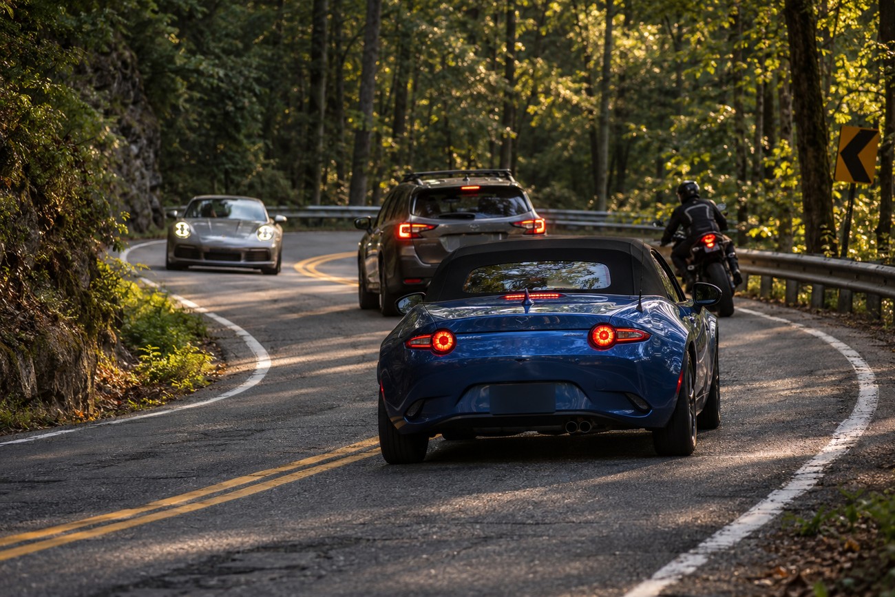 A blue Mazda Miata driving on a winding forest road with other vehicles ahead