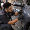 Mechanic in safety glasses and black gloves measuring panel gaps on a gray car door in a workshop