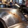 A mechanic wearing safety glasses and gloves inspects surface rust on a metal panel with a flashlight