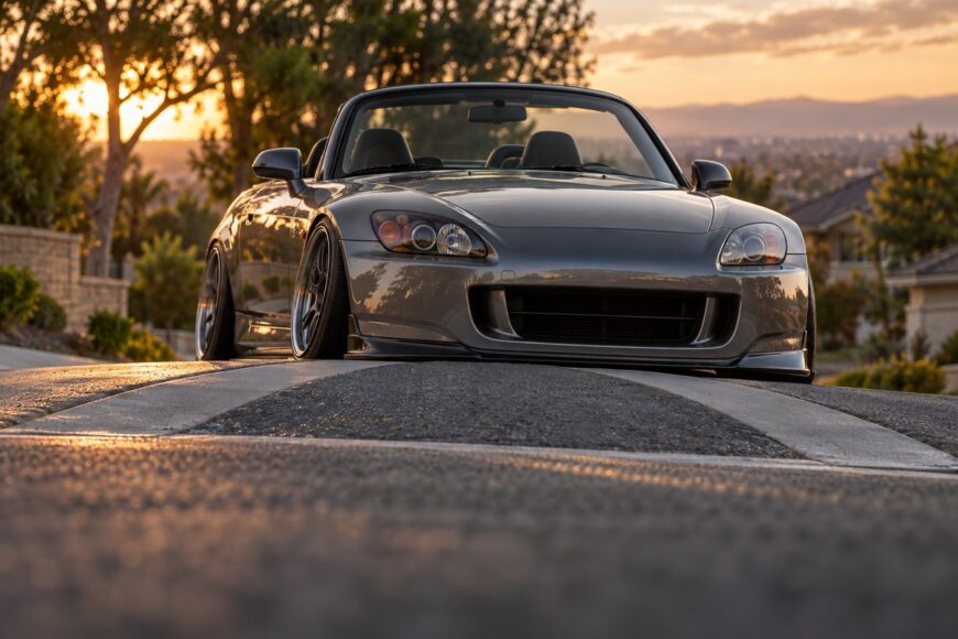 A lowered gray Honda S2000 convertible stuck on a humped road crossing at sunset
