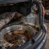 Severely rusted spare tire well and trunk floor inside an open car trunk with water damage