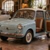 A light blue and white Autobianchi Bianchina vintage car parked in a workshop with open doors and brown interior