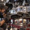 Mechanic wearing safety glasses and gloves uses a flashlight and a ruler to inspect damaged frame rail of a car on a frame machine
